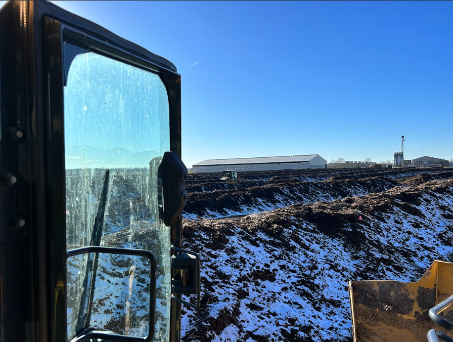 View from inside heavy machinery cab, looking out at a snowy construction site with a building and clear blue sky.
