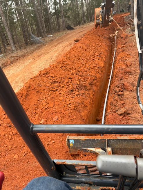 Trench dug with an excavator, with a white pipe installed. Red dirt road in a wooded area.