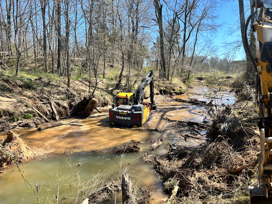 A yellow excavator clearing debris from a muddy creek in a wooded area.