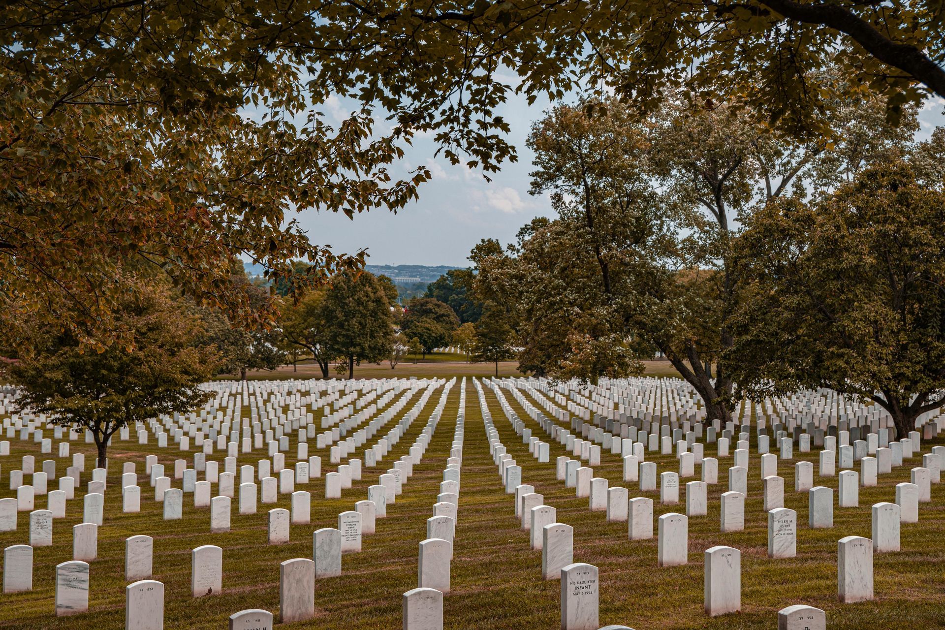 Rows of white headstones in a cemetery, under trees and a partly cloudy sky.