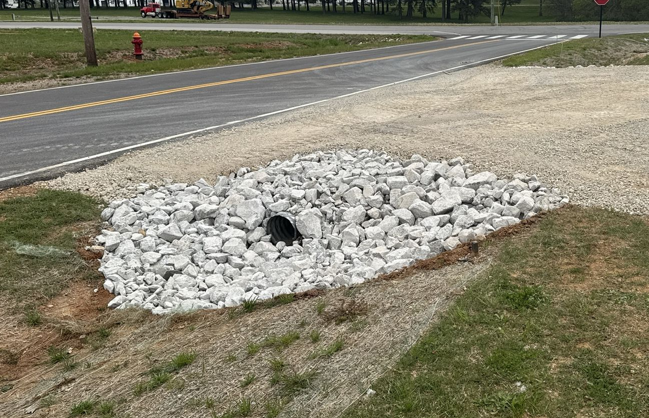 Storm drain covered in rocks next to a road and grassy area.