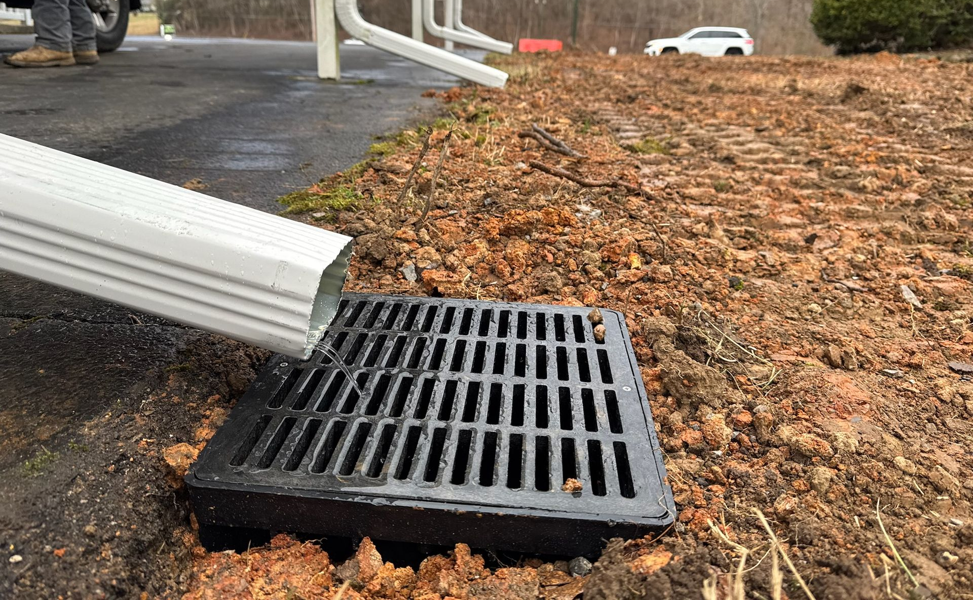 Downspout draining into a black grate on the edge of a driveway, surrounded by dirt.