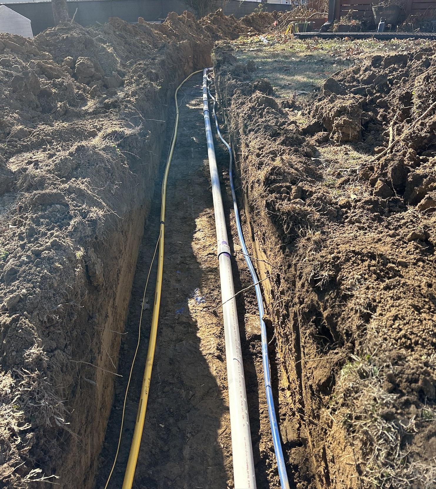 Trench with buried utility lines: yellow gas, white pipe, and black electrical cable. Soil fills the trench.