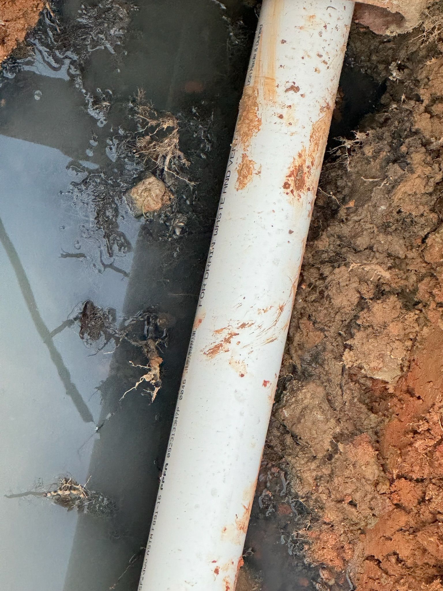 White pipe in a trench with dirty water and reddish-brown soil.