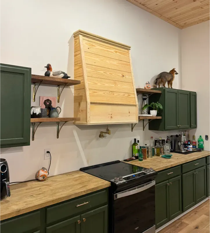 Green kitchen cabinets with wooden countertops, range, and a light wood range hood.