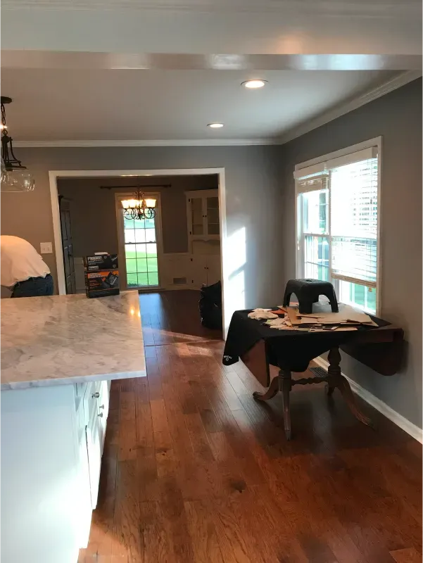 Hardwood-floored dining room with a table near a window. Gray walls, white trim, and a view into another room.