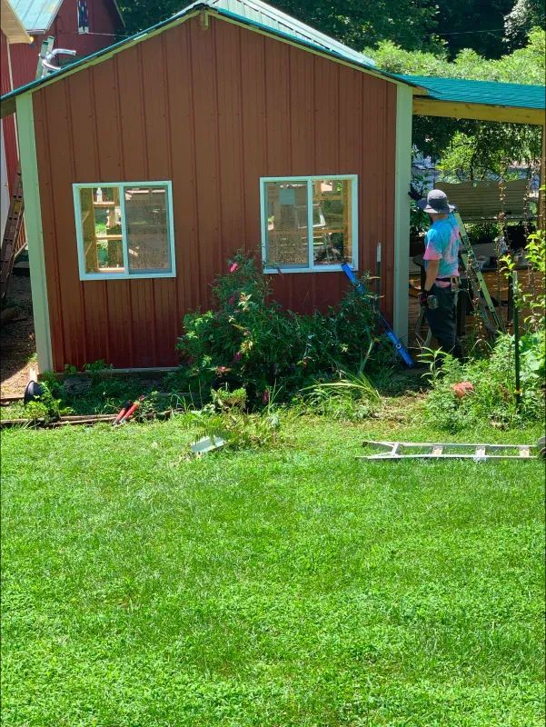 Red shed with white-framed windows, surrounded by greenery and a lawn. A person works near the shed.
