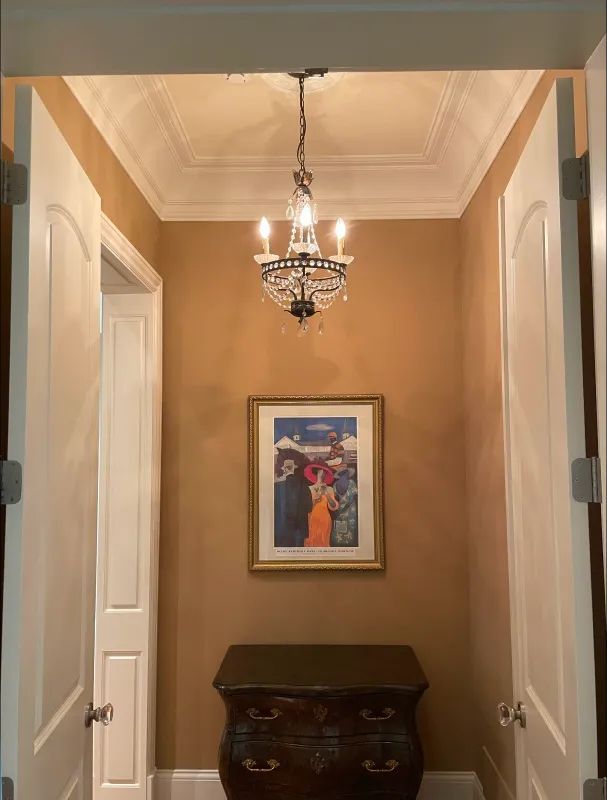Hallway with gold-colored walls, ornate ceiling, chandelier, artwork, and dark wooden chest between white doors.