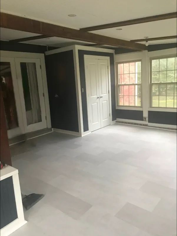 Empty room with light gray floor, dark blue walls, white doors/trim, and windows. Dark wood beams cross the white ceiling.