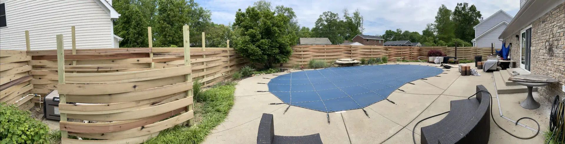 Backyard with a pool covered by a blue tarp, surrounded by a fence and stone patio.