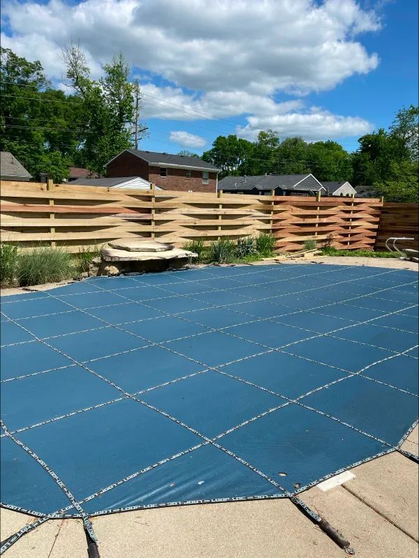 Pool covered with blue tarp, surrounded by stone deck, wooden fence, under a blue sky.