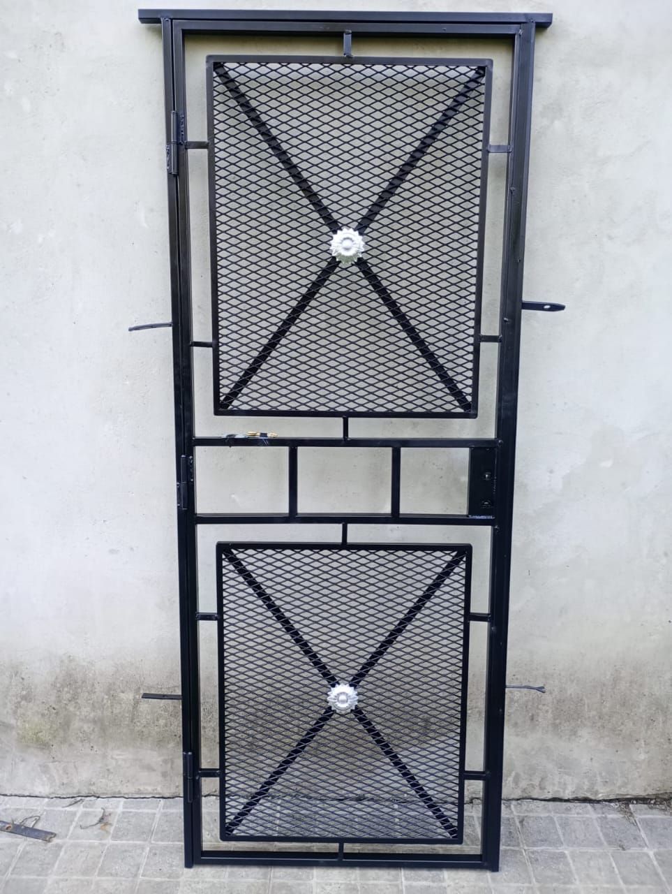 Puerta de seguridad de metal negro con paneles de malla y herrajes contra una pared gris claro.
