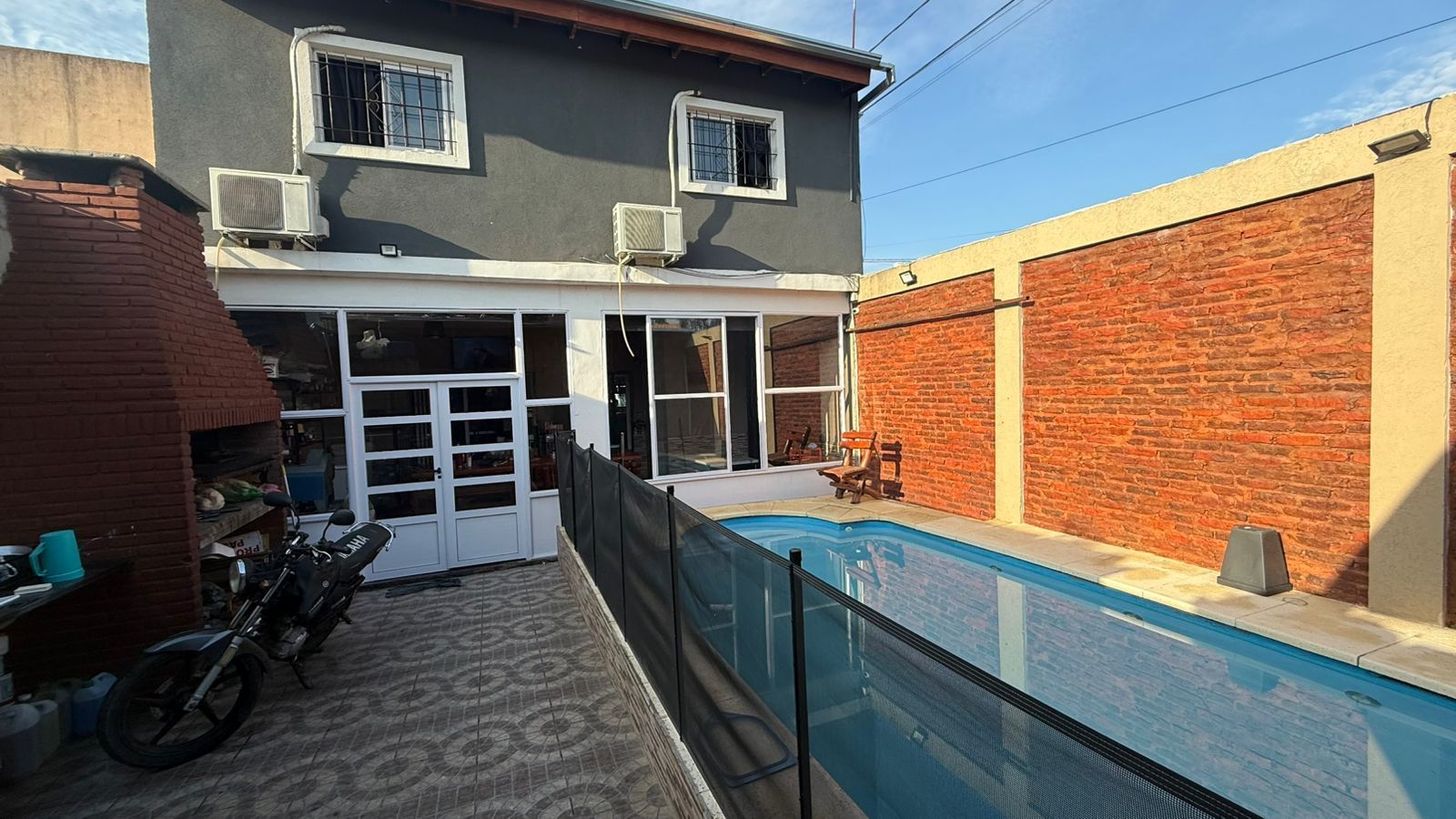 Patio trasero con piscina, casa gris, pared de ladrillo, motocicleta, asientos al aire libre.