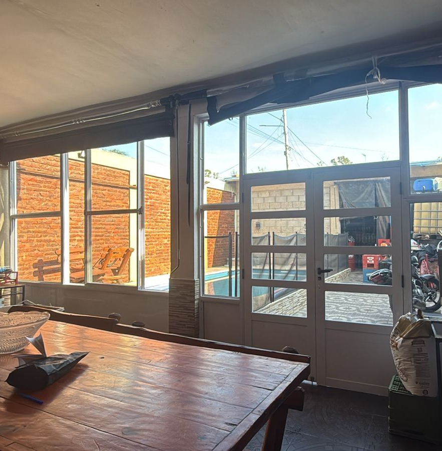 Vista interior: mesa de madera, ventanas, pared de ladrillo, puertas a zona de piscina, soleado.