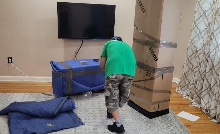 A man is standing in a living room surrounded by boxes and a television.