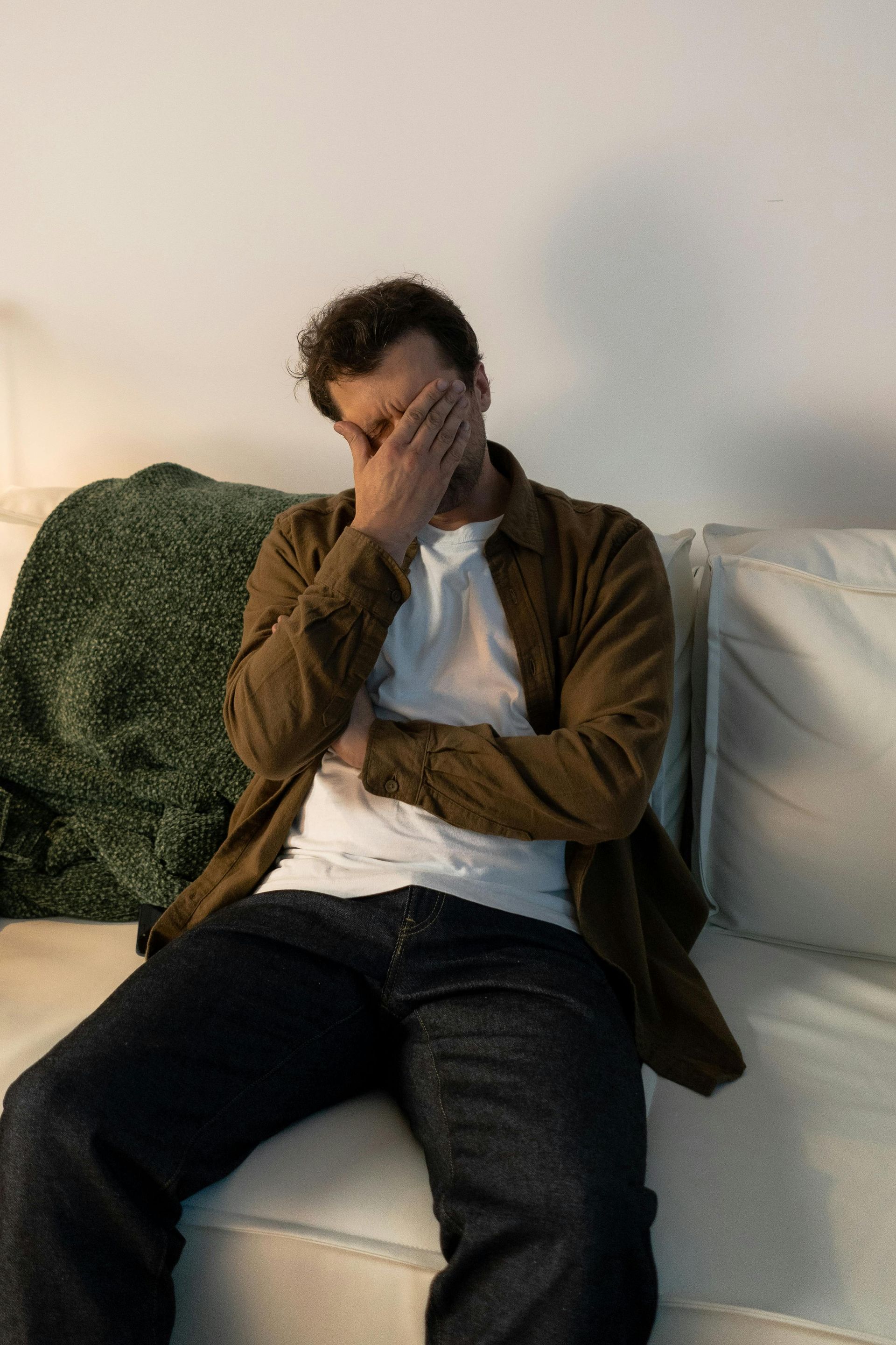 Man sitting on a white couch, face in hand, wearing a brown shirt and jeans.