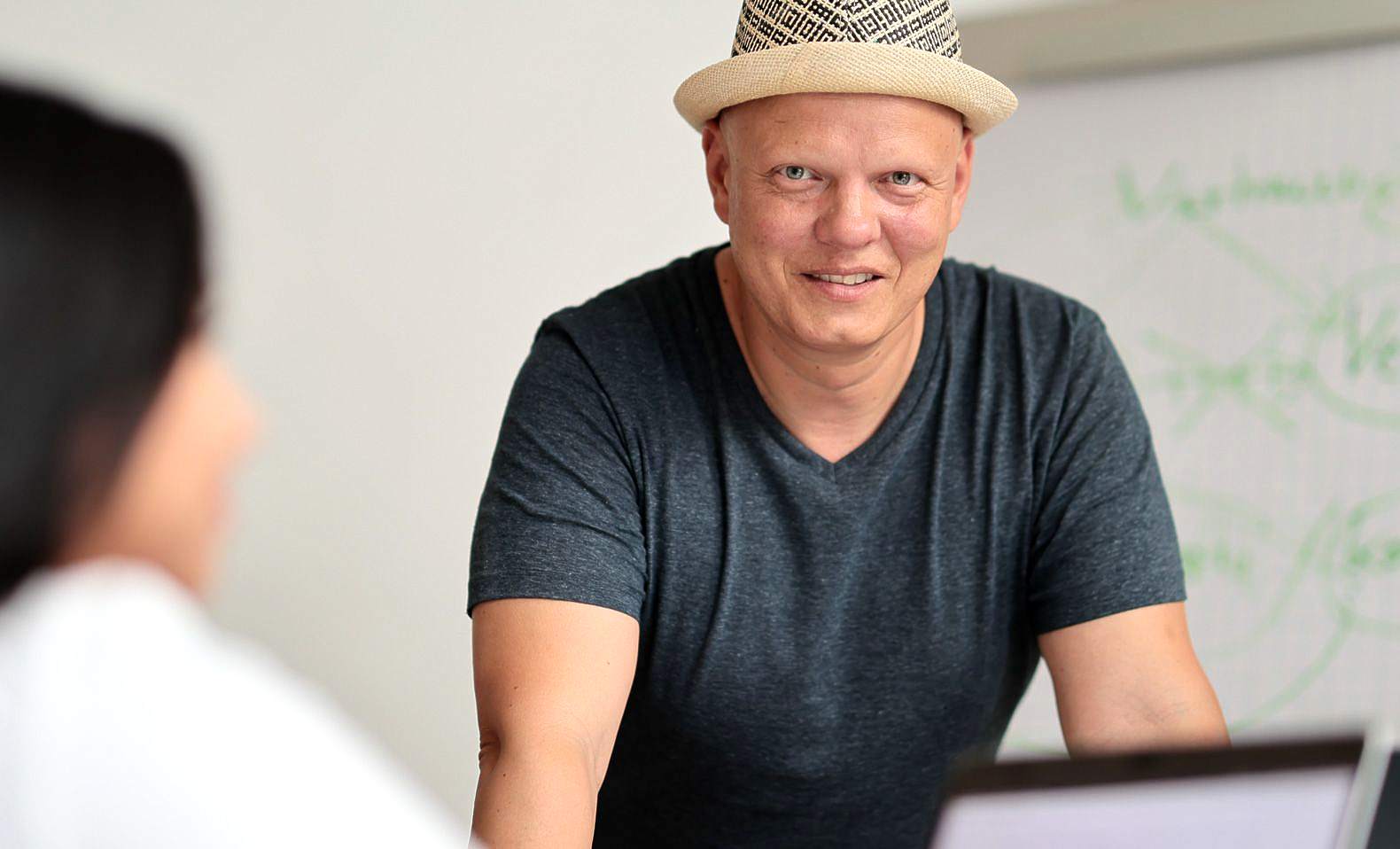 Daniel Docter mit Hut lächelt eine Person an, die sich in einem Büro über einen Tisch beugt, im Hintergrund ist ein Whiteboard zu sehen.