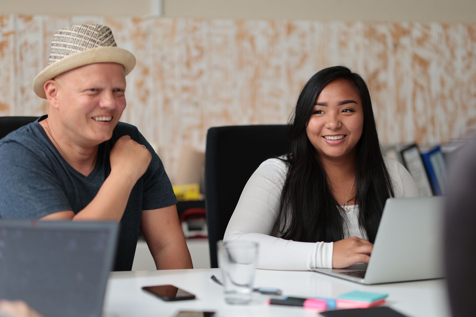 Mann mit Hut und Frau lächelnd an einem Tisch mit Laptops und einem Glas Wasser.