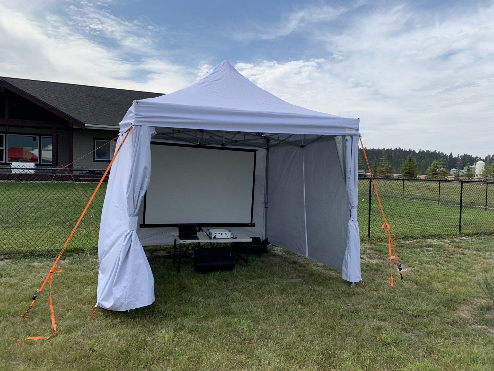 A white tent with a projector screen attached to it is sitting in the grass in front of a house.