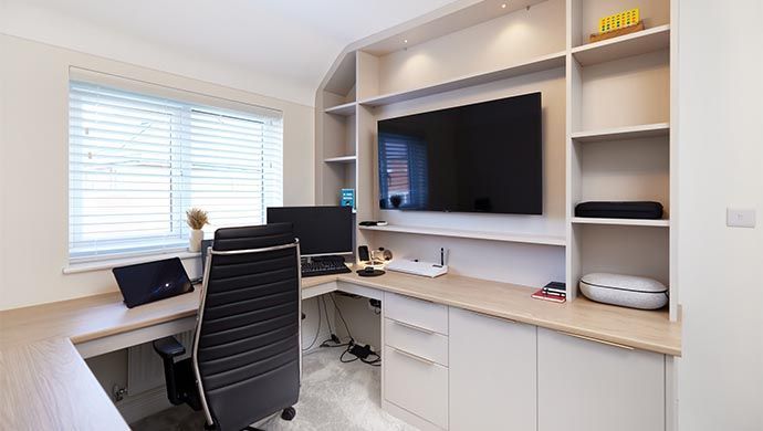 AD Construction NJ - Home office with built-in desk, shelving, TV, and monitor. Light-colored wood and neutral walls.