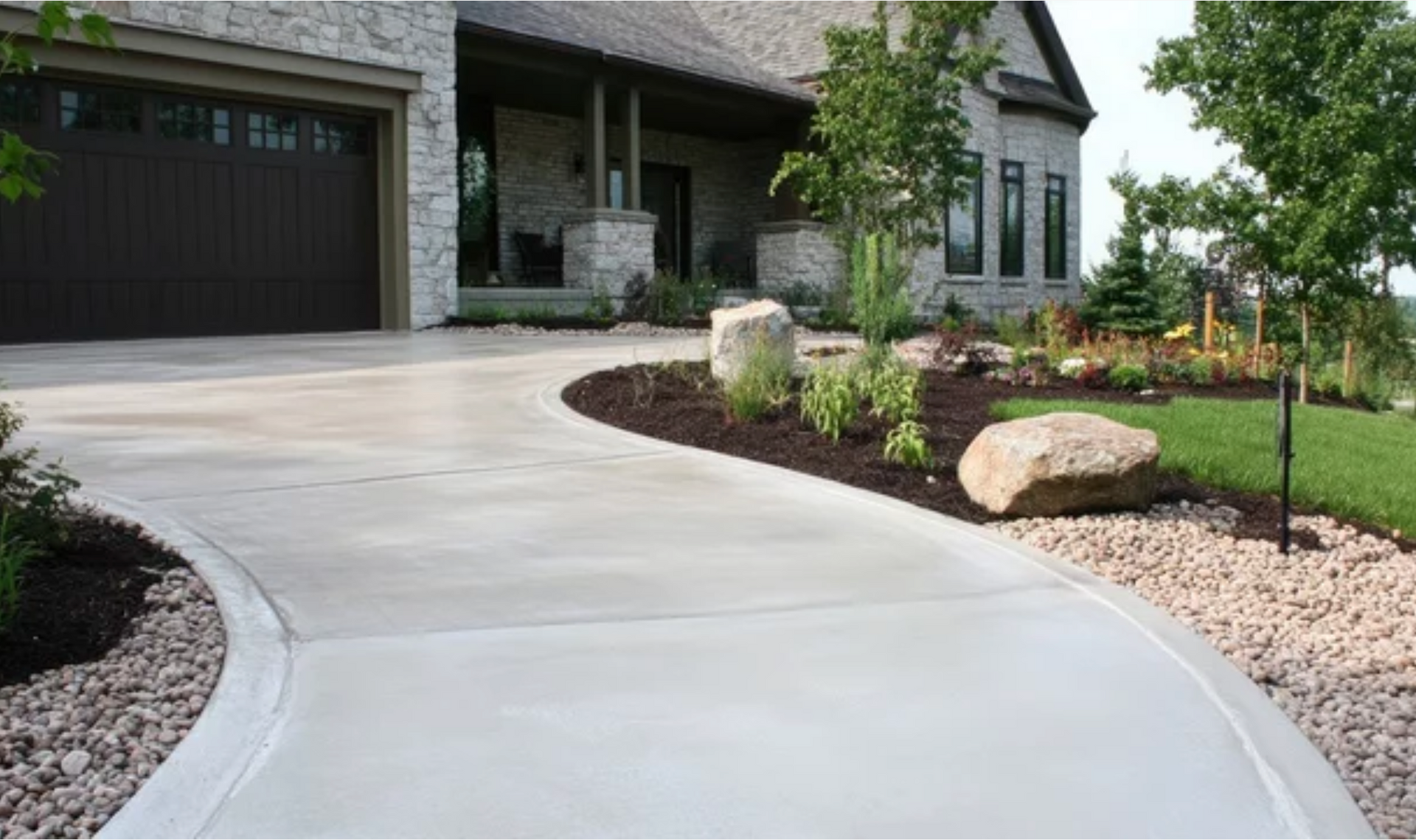 AD Construction NJ - Concrete driveway curves towards a stone house, lined with landscaping and a dark garage door.
