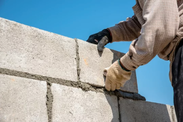 AD Construction NJ - Hands of a construction worker laying gray concrete blocks, against a blue sky.