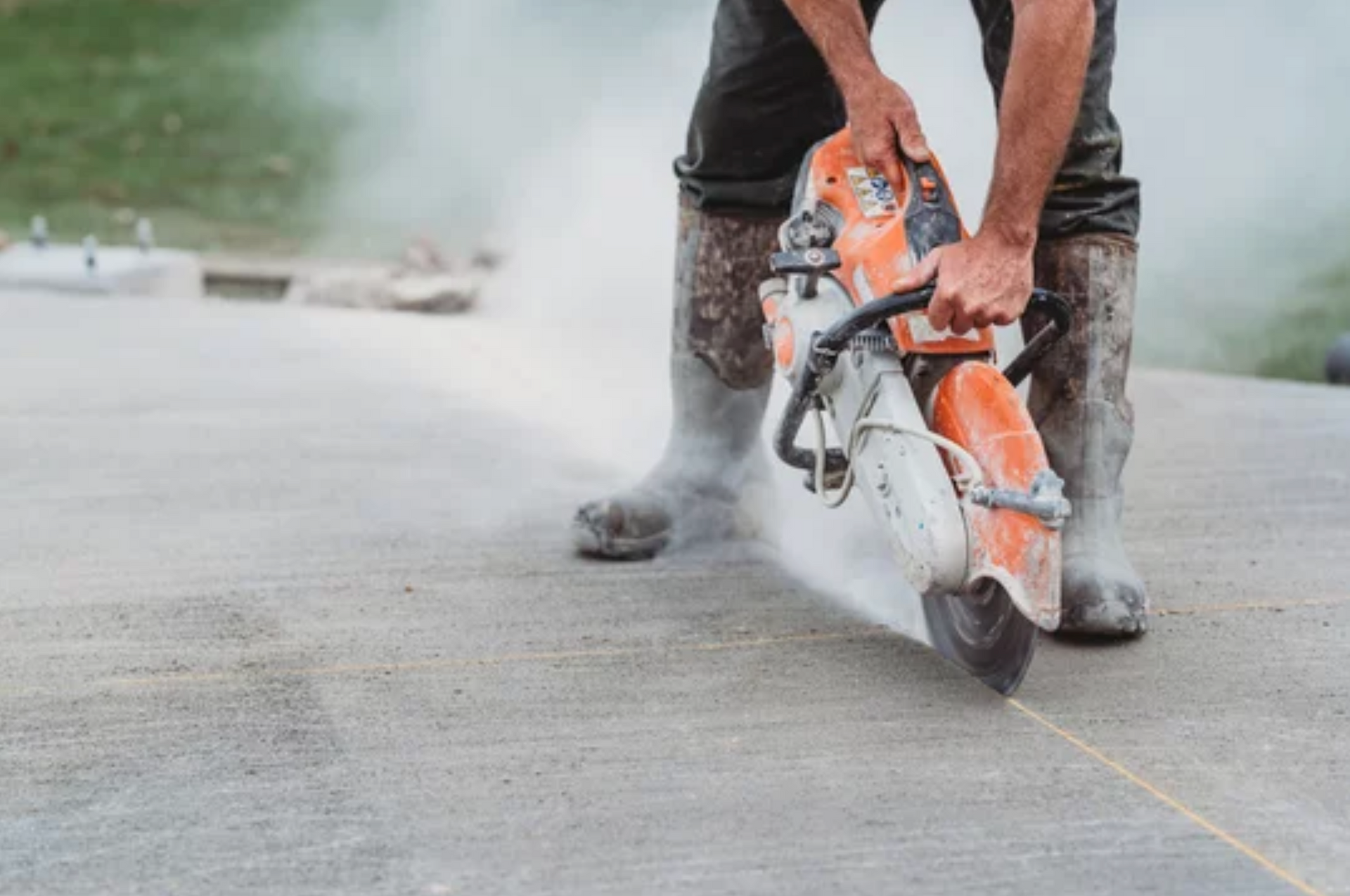 AD Construction NJ - Person in boots uses a concrete saw, creating dust and a cutting line on a concrete surface outdoors.
