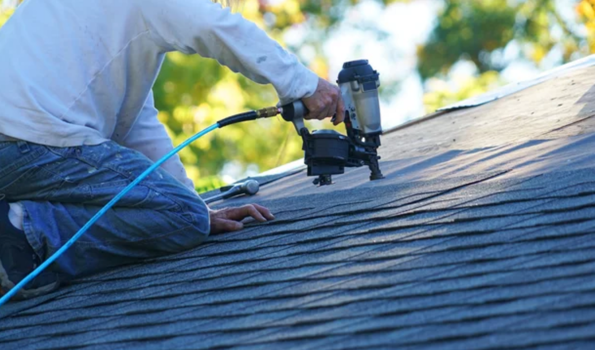 AD Construction NJ - Roofer kneeling on a shingled roof, using a nail gun; blue hose connected.