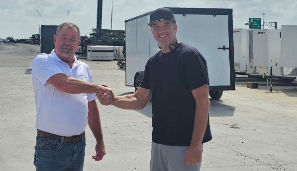 Two people shaking hands outdoors in a trailer sales lot with white cargo trailers parked in the background.
