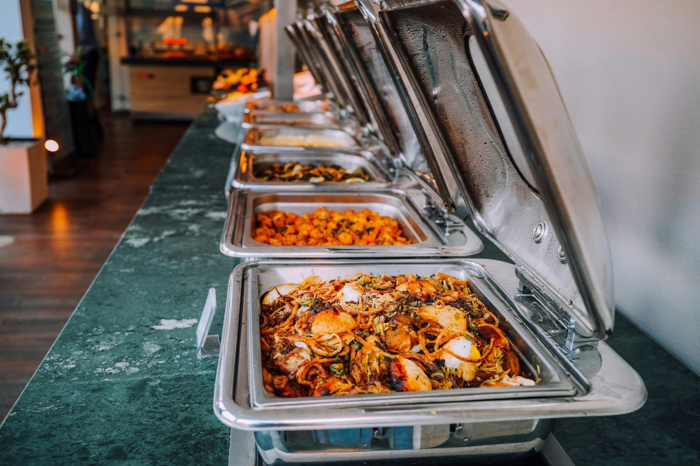 Buffet line with various dishes in silver chafing dishes, on a long, green-topped table.