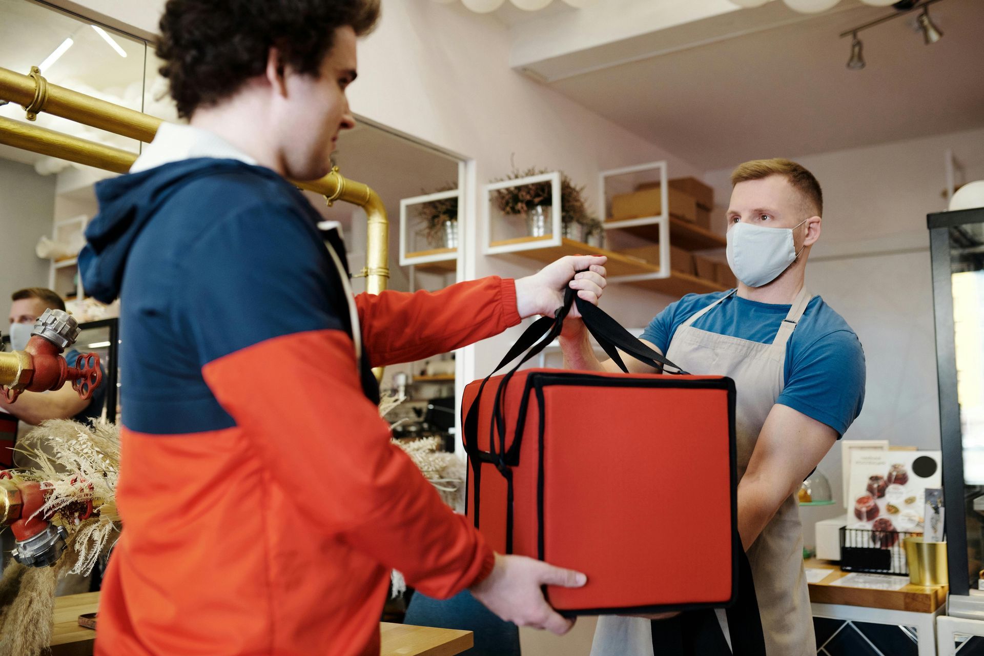 Man receiving red delivery bag from masked worker in shop.