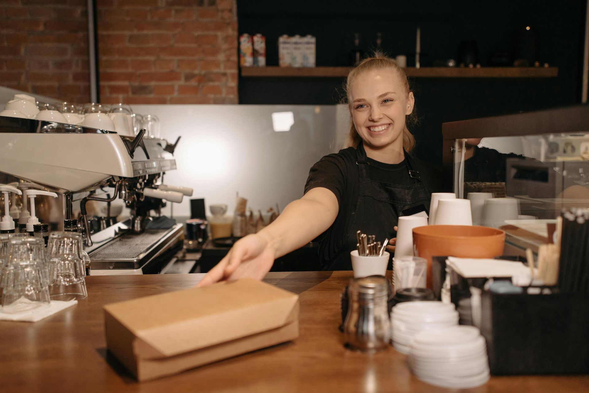 Barista smiling, handing a brown food box across a counter in a cafe.