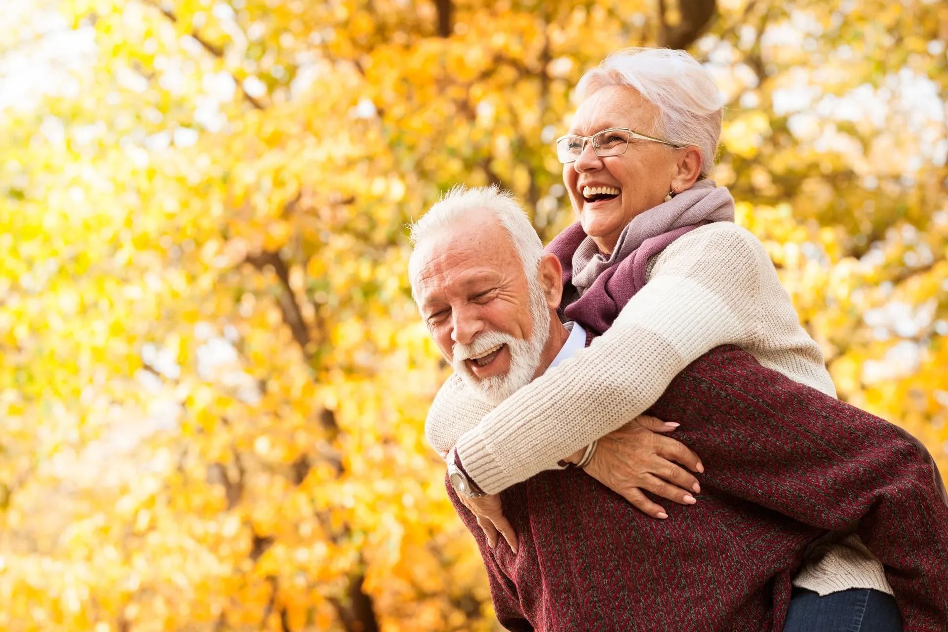 An older man is giving an older woman a piggyback ride in a park.