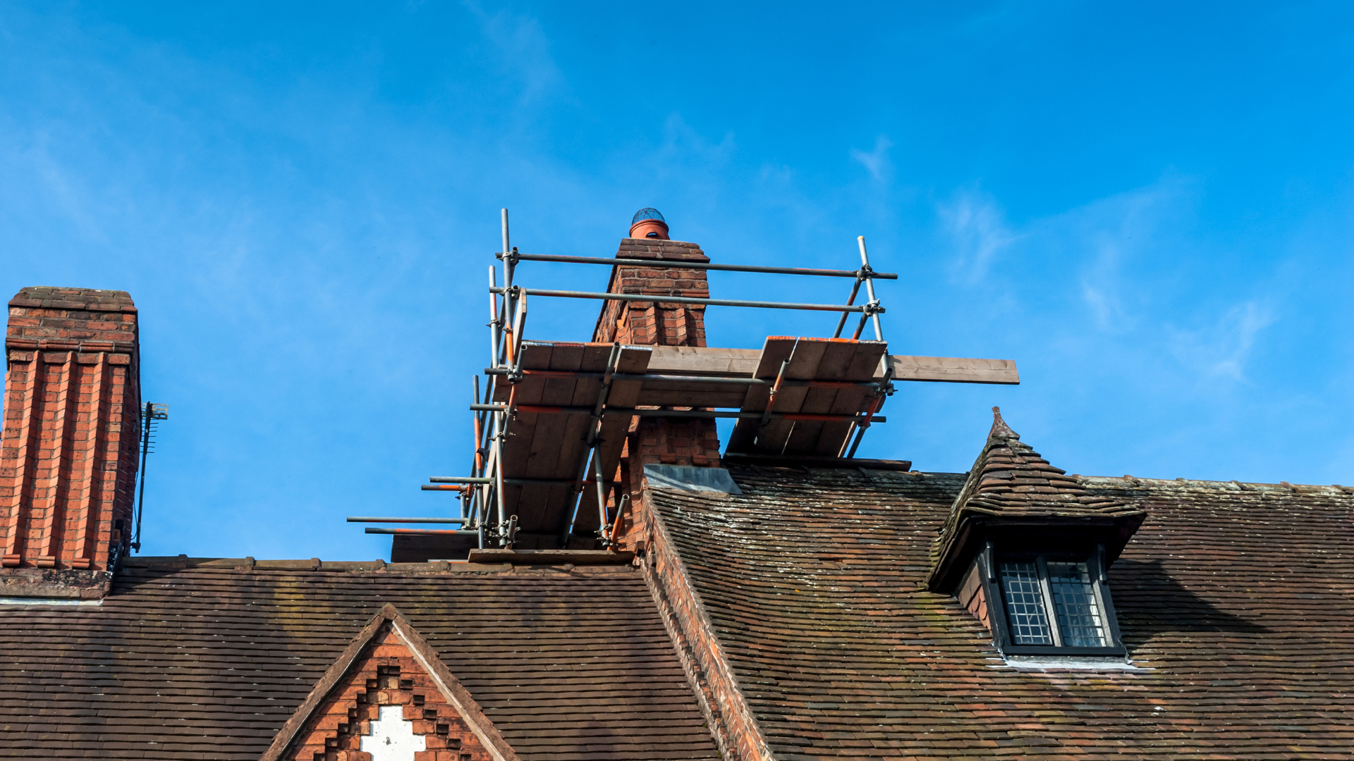 Chimney repair: Scaffolding surrounds a brick chimney on a rooftop with a clear blue sky background.