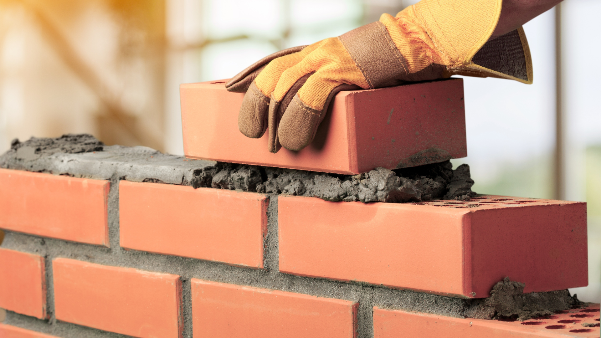 Bricklayer laying a red brick onto mortar on a brick wall, wearing a brown and yellow glove.