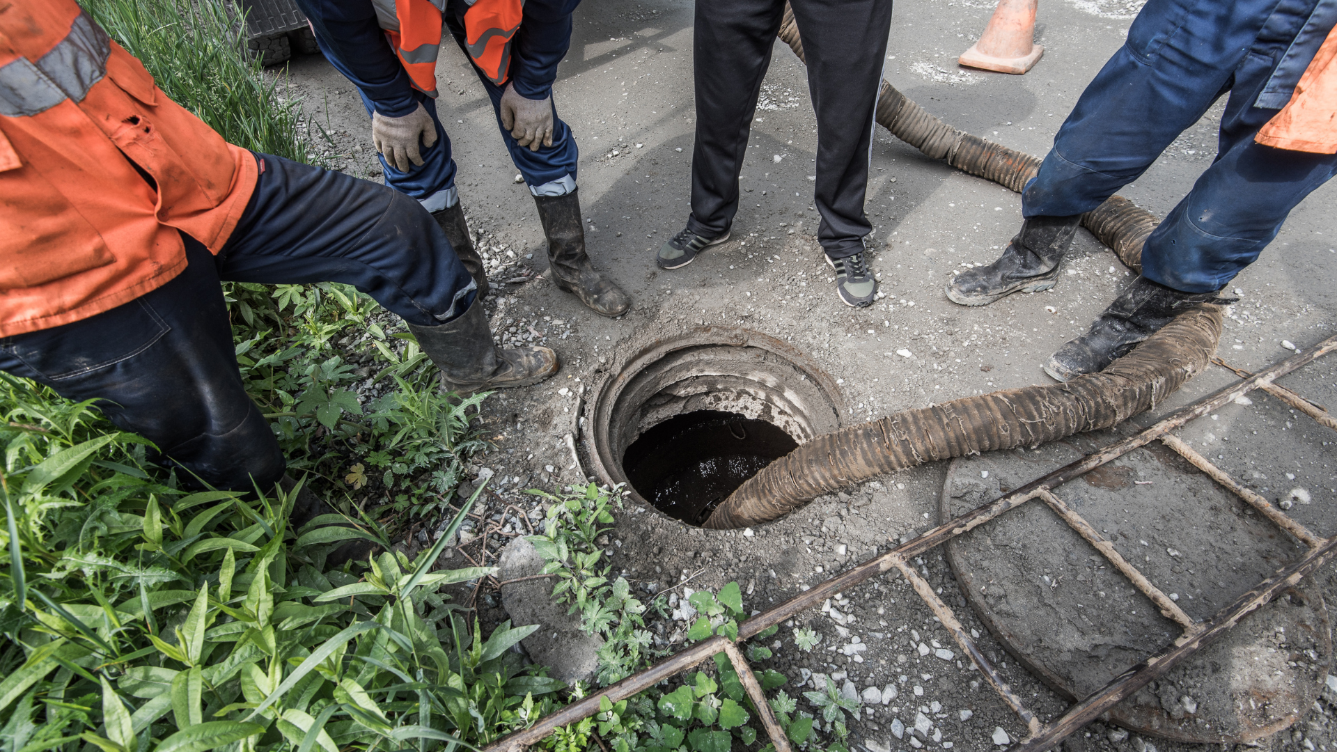 Workers around an open manhole on a road, with hose and green grass.