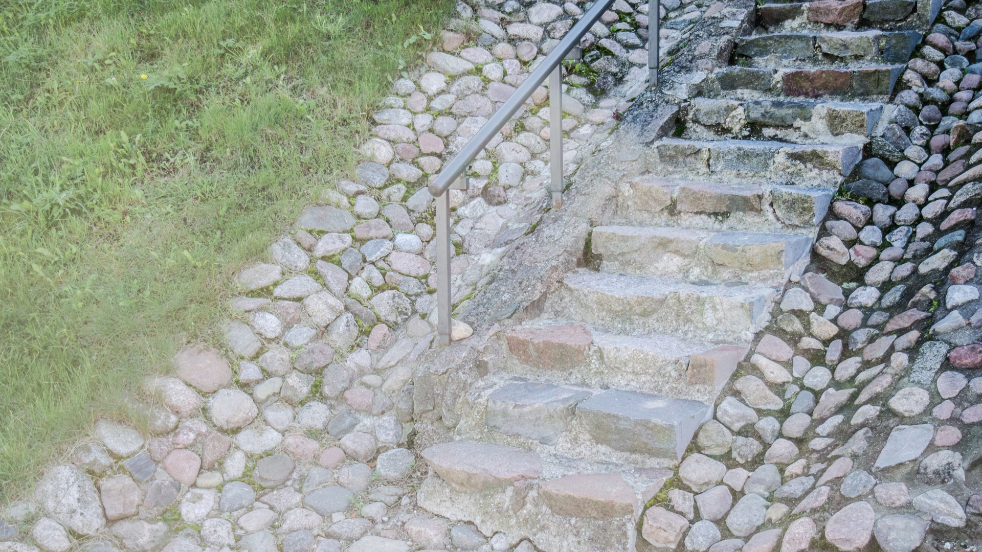 Stone steps with metal handrail built into a stone wall, beside grass.