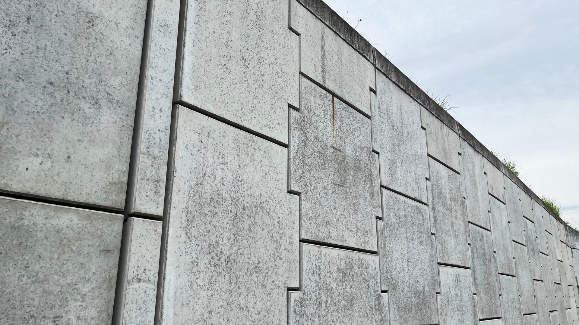 Gray concrete retaining wall with geometric block patterns against a cloudy sky.