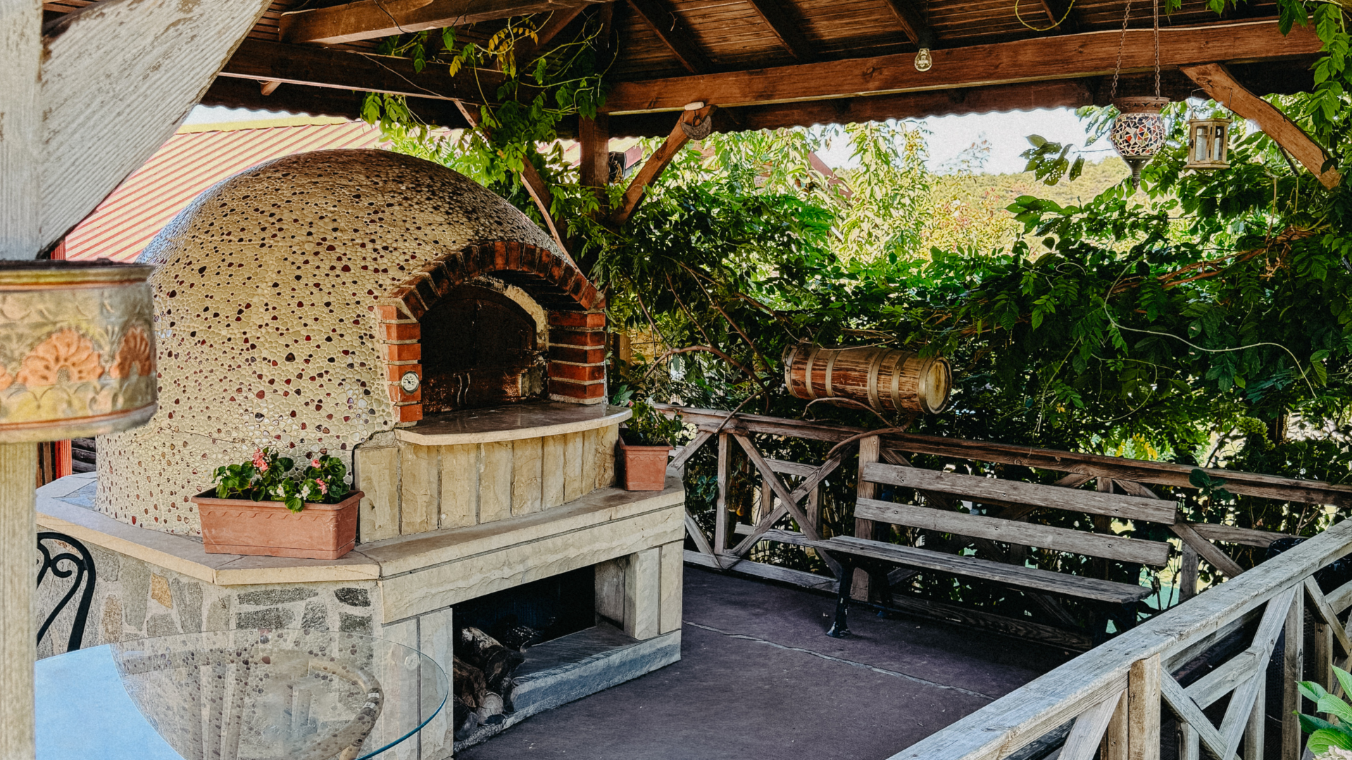 Pizza oven on patio with bench, wooden fence, and greenery.