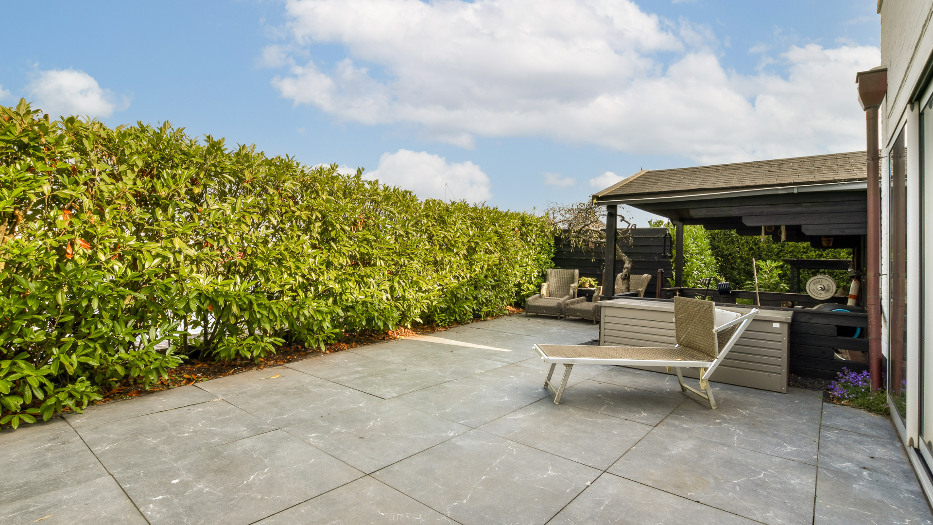 Patio with gray tiles, a green hedge, and lounge chairs under a shaded area.