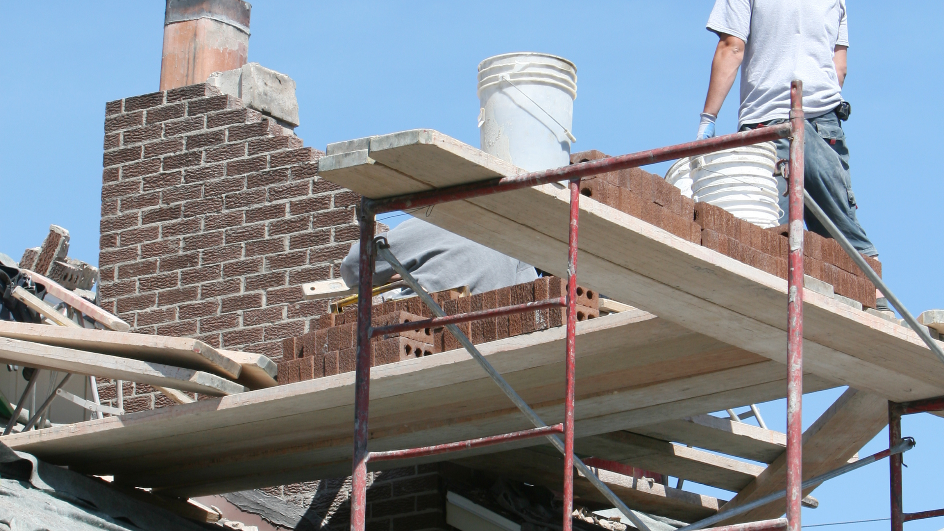 Man rebuilding a brick chimney on a rooftop, using scaffolding and buckets.