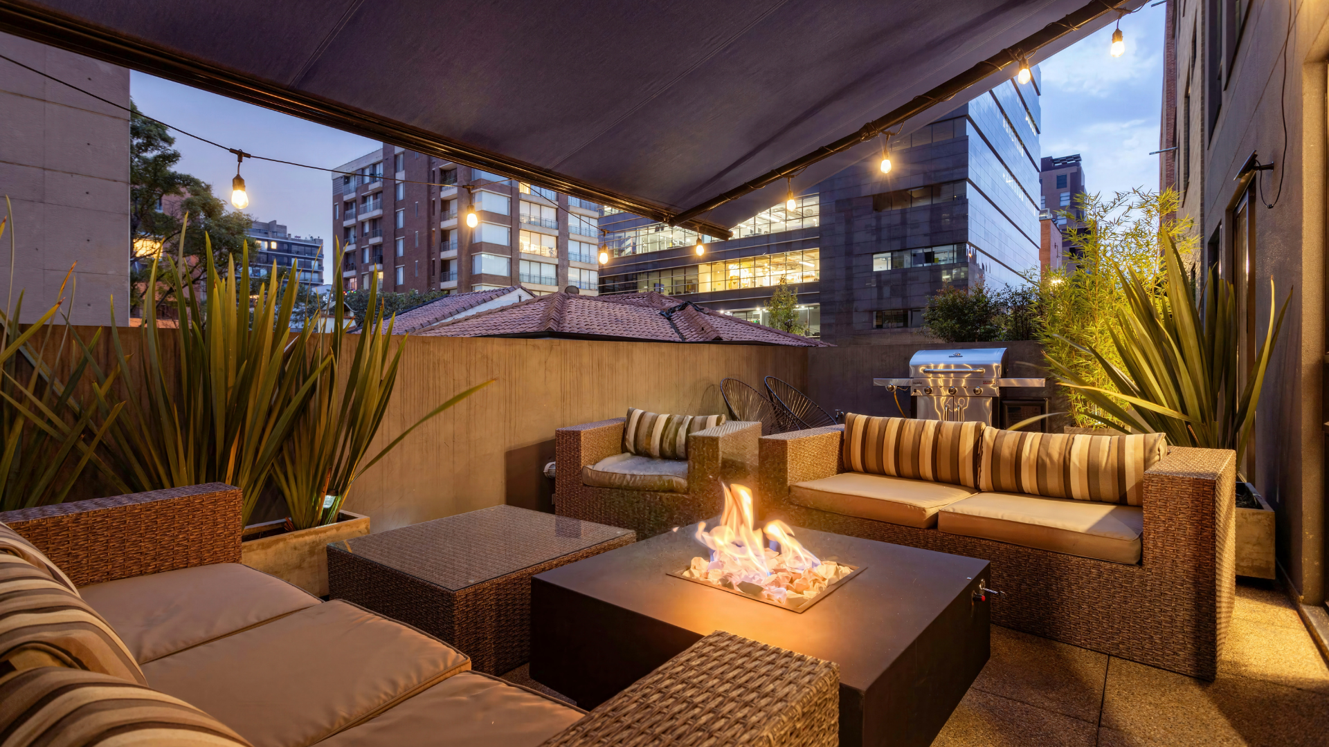 Outdoor patio with seating, fire pit, and string lights, with city buildings in background.