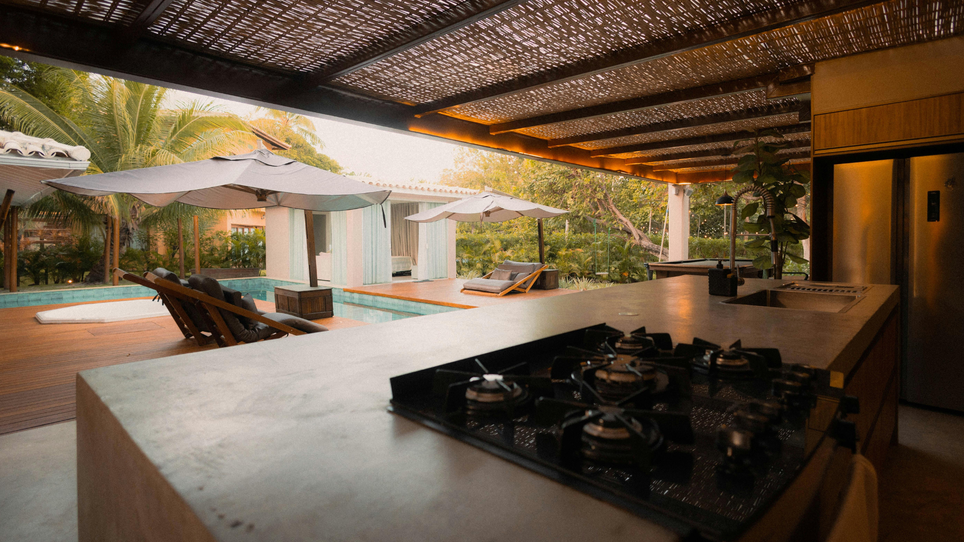 Outdoor kitchen with a stove, overlooking a pool with umbrellas and lounge chairs.