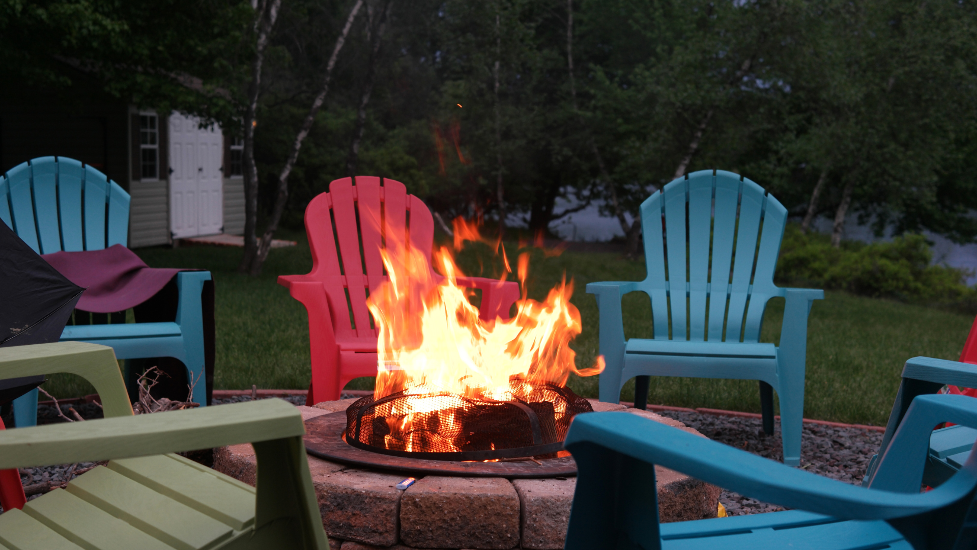 Fire pit with large flames, surrounded by colorful Adirondack chairs on a lawn near a small building.