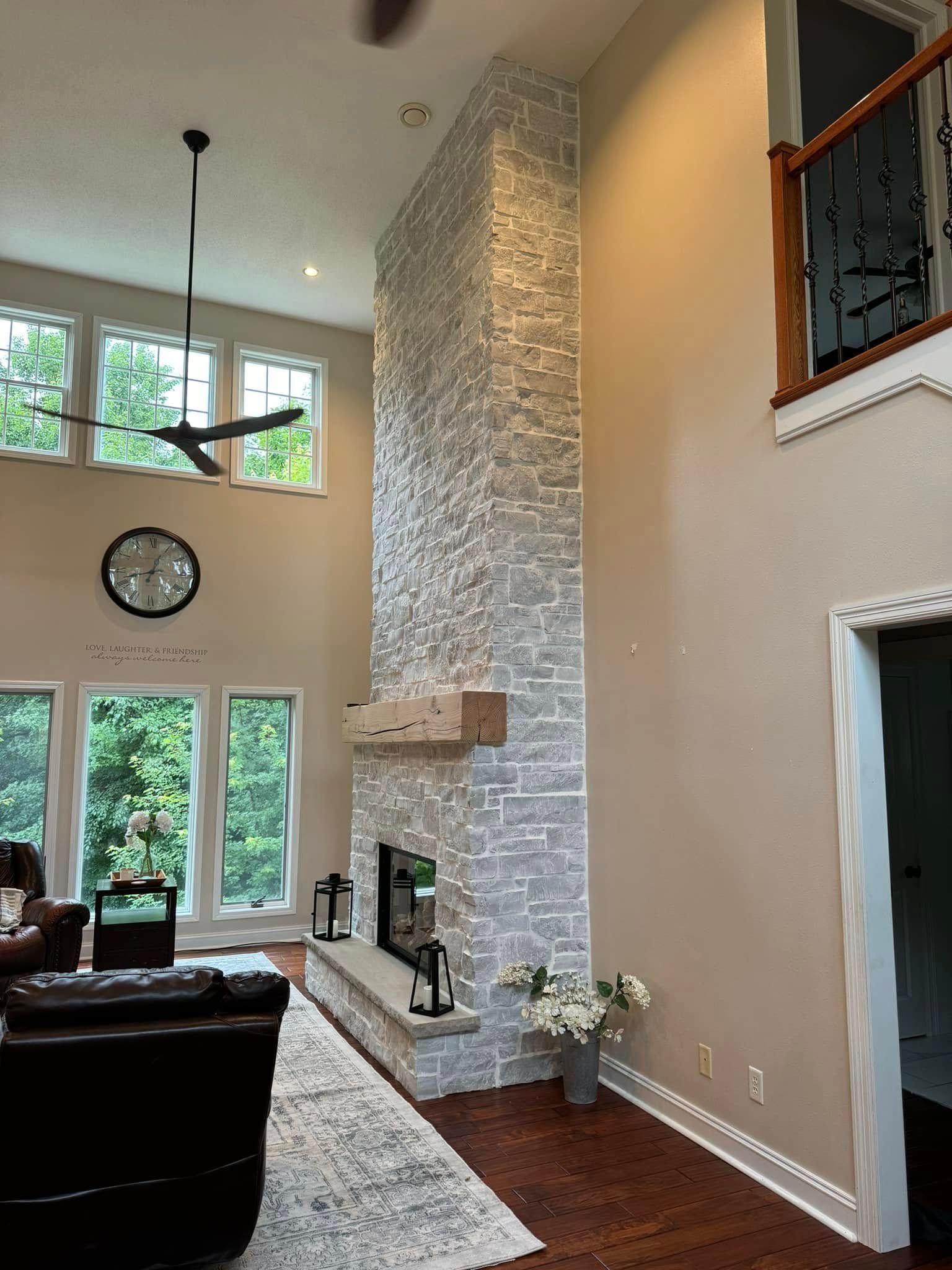 A tall stone fireplace in a two-story room, wooden floors, windows with outdoor views.