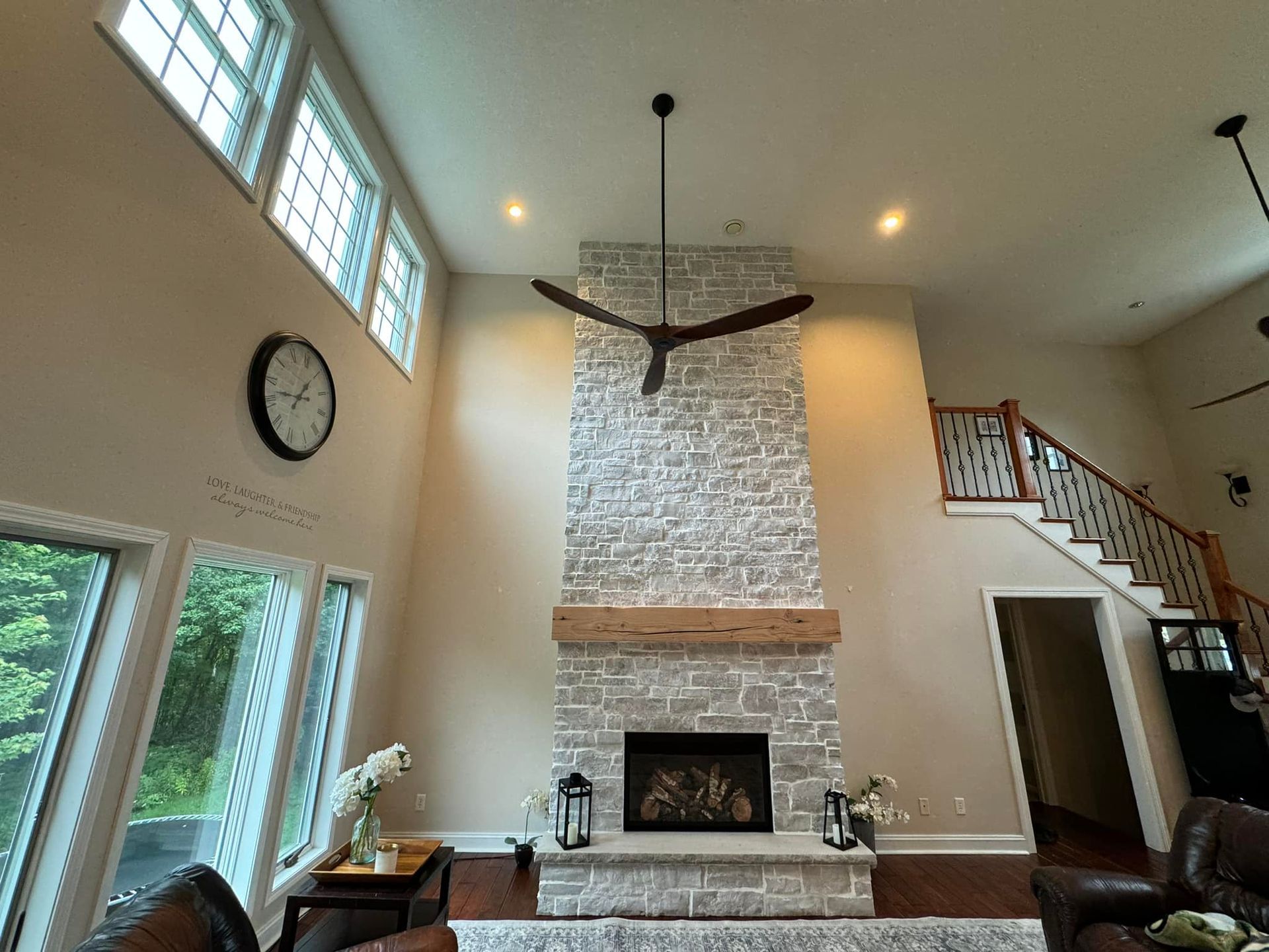 Living room with a stone fireplace, dark ceiling fan, tall windows, and a staircase.