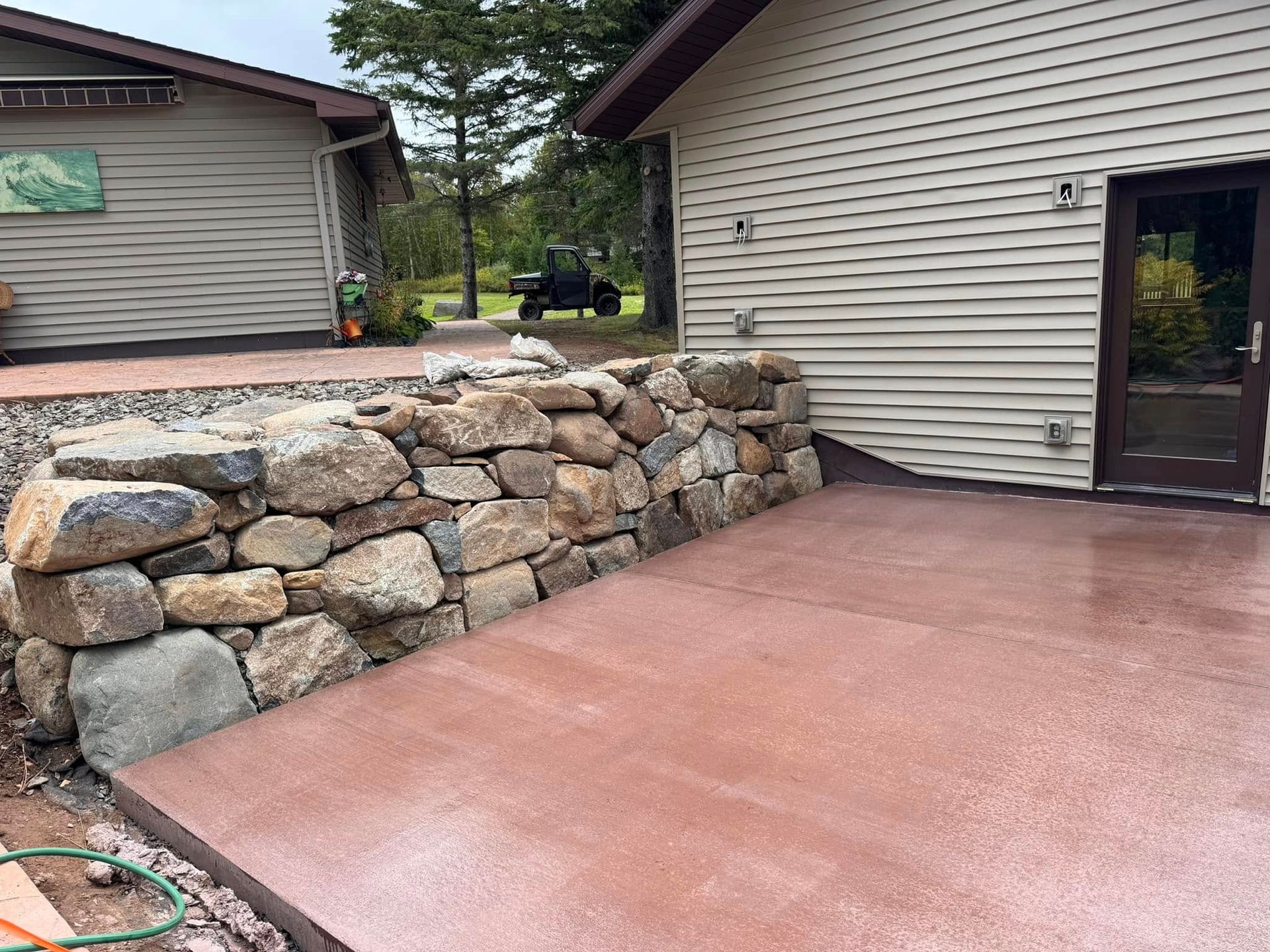 Red concrete patio next to a stone retaining wall and a building with tan siding and a dark brown door.