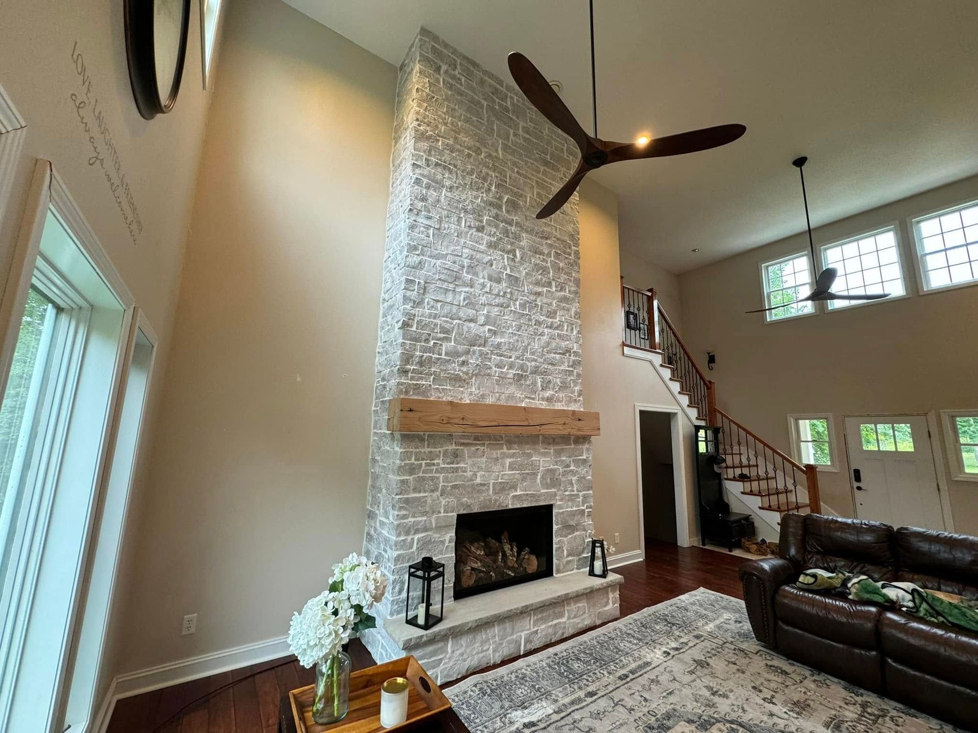 Living room with stone fireplace, wooden mantel, and high ceilings. Brown leather couch and ceiling fans.