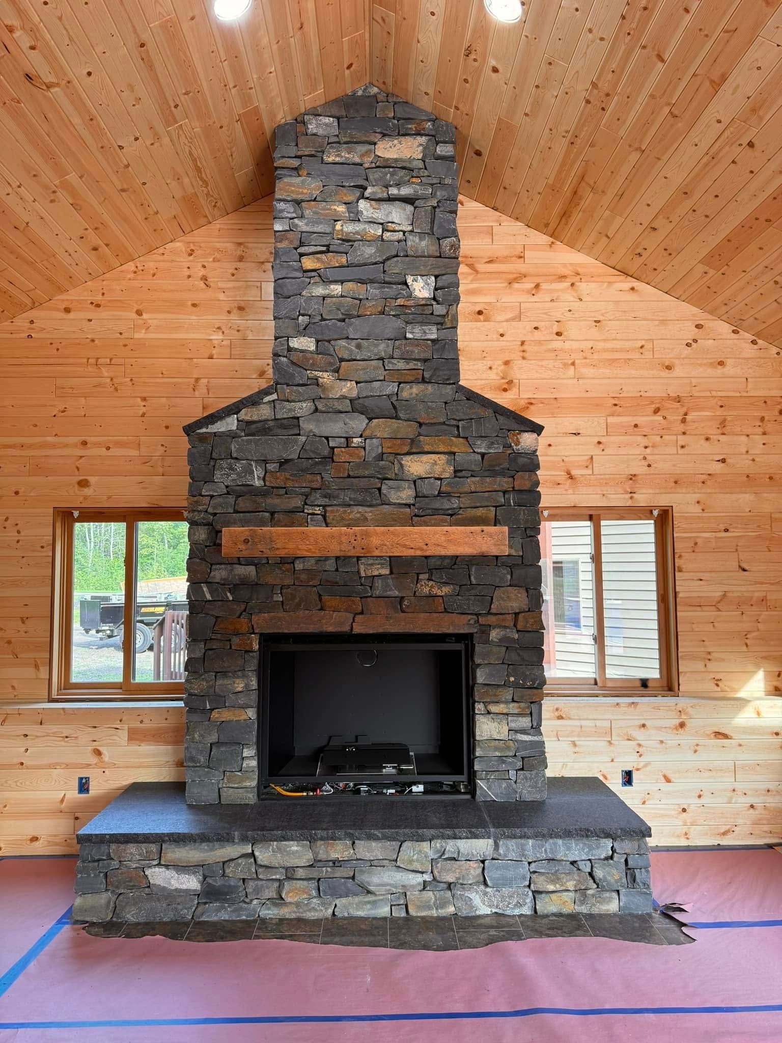 Stone fireplace in a wood-paneled room. Firebox has a black interior. Two windows flank the fireplace.