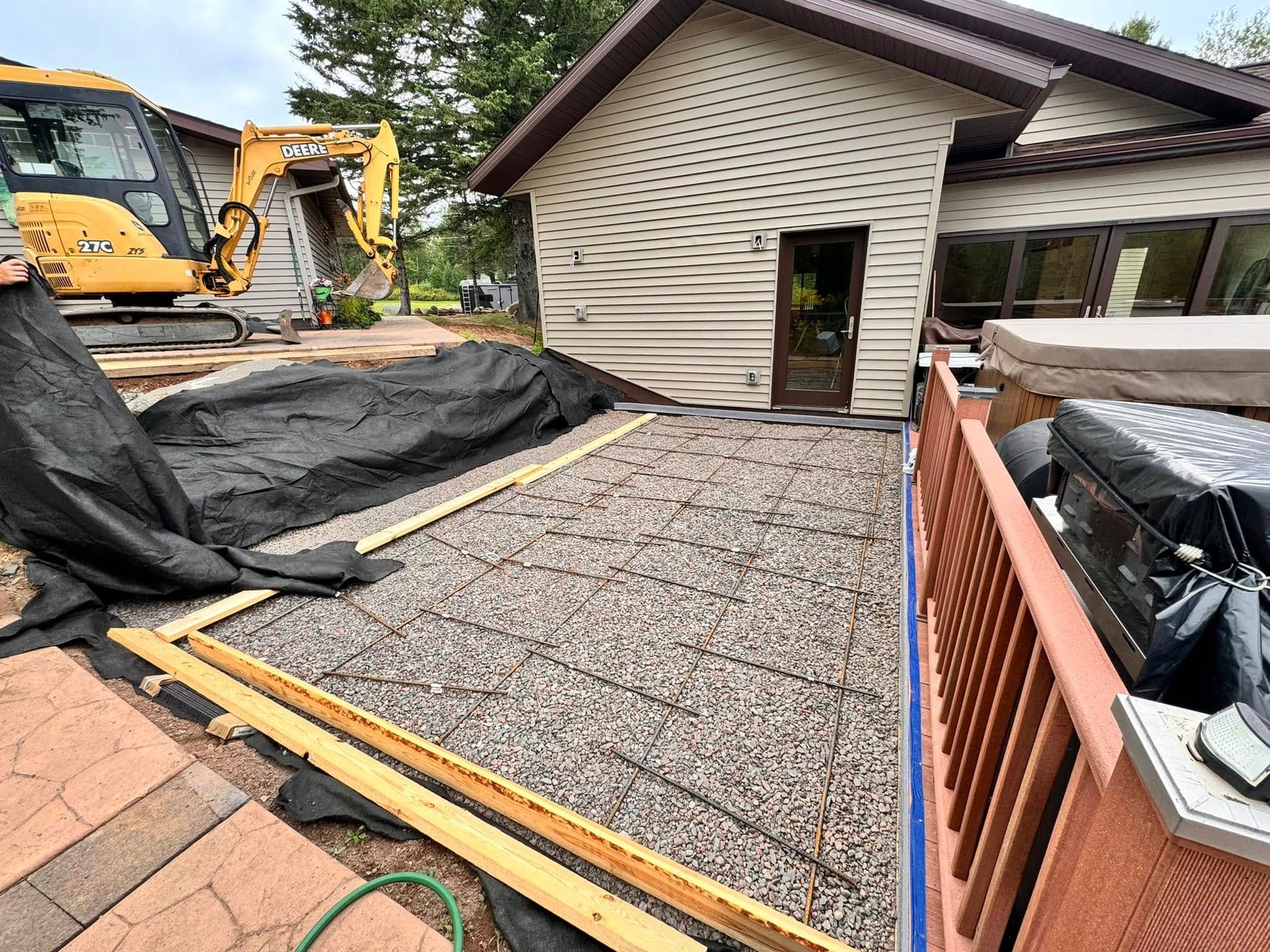 Patio construction site: gravel base, wooden frame, rebar grid, black tarp, excavator in background.